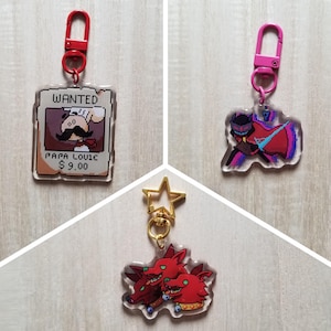 Puede incluir: Tres llaveros acrílicos con diferentes diseños. Uno presenta un cartel de "Se busca" con "Papa Louie" y un precio. Otro muestra un personaje pixelado con capa. El tercer llavero tiene tres criaturas rojas con aspecto de perro.