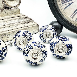 Op de afbeelding: Witte keramische kastknoppen met een blauw bloemmotief en zilverkleurige hardware. De knoppen zijn rond en hebben een decoratief patroon. De afbeelding bevat ook een klok en een decoratieve houten basis.
