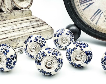 May include: White ceramic cabinet knobs with a blue floral design and silver hardware. The knobs are round and have a decorative pattern. The image also includes a clock and a decorative wooden base.