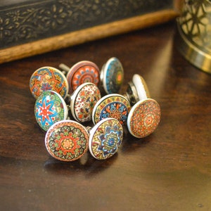 May include: A collection of ten colourful ceramic cabinet knobs with intricate patterns in shades of blue, red, orange, green, and white. The knobs are arranged in a circle on a dark brown wooden surface.