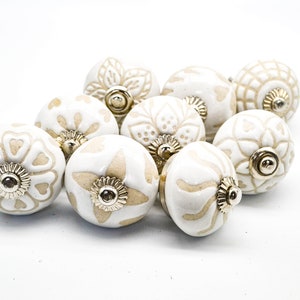 Op de afbeelding: Tien witte keramische kastknoppen met een beige bloemenpatroon. Elke knop heeft een gouden metalen basis.