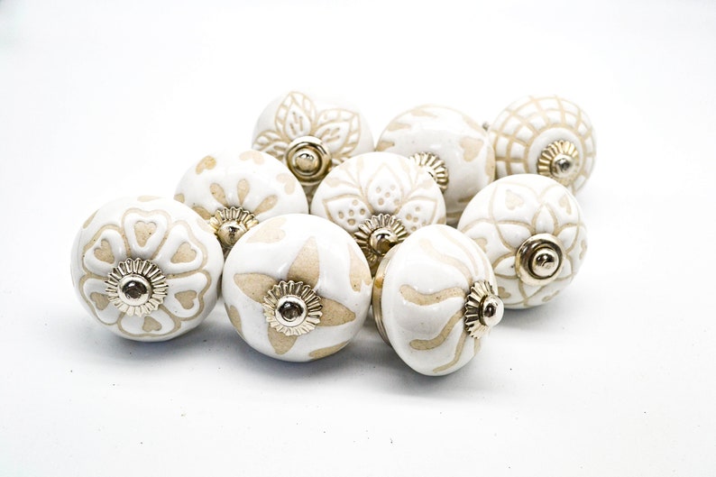 Op de afbeelding: Tien witte keramische kastknoppen met een beige bloemenpatroon. Elke knop heeft een gouden metalen basis.