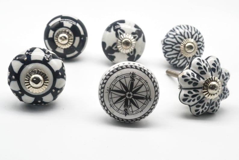 K&ouml;nnte beinhalten: Sechs schwarze und wei&szlig;e Keramik-Schrankkn&ouml;pfe mit verschiedenen Designs. Die Kn&ouml;pfe zeigen einen Kompass, geometrische Muster und florale Motive.