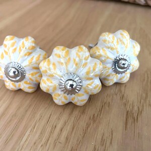 May include: Three floral cabinet knobs. Each knob is white with yellow leaf patterns and a silver metal centre. The knobs are arranged on a light brown wooden surface.