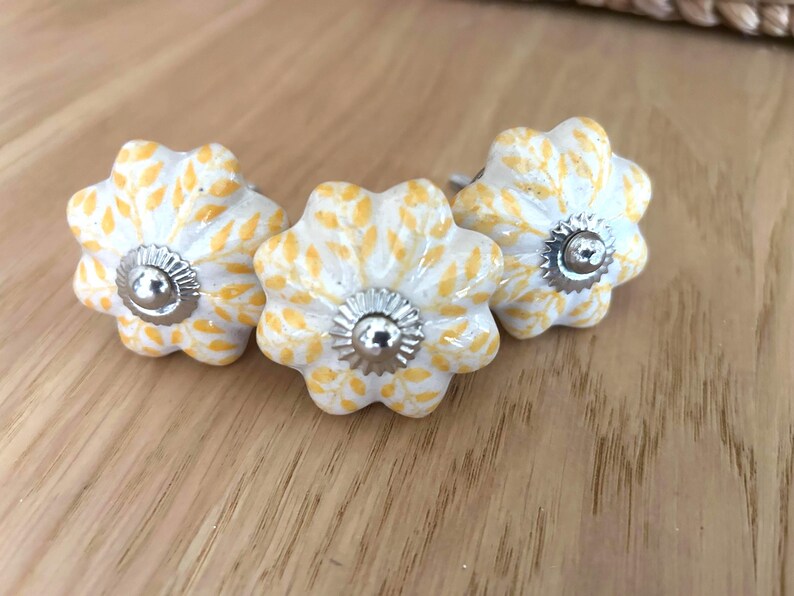 May include: Three floral cabinet knobs. Each knob is white with yellow leaf patterns and a silver metal centre. The knobs are arranged on a light brown wooden surface.