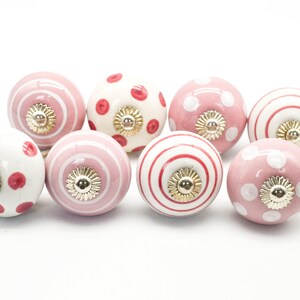 May include: Set of eight pink and white ceramic cabinet knobs with different patterns, including polka dots, stripes, and swirls. Each knob has a gold-colored accent.
