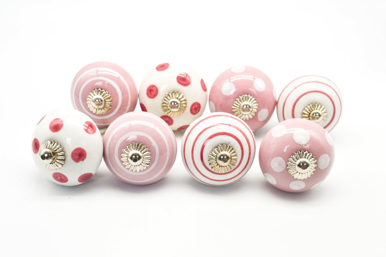May include: Set of eight pink and white ceramic cabinet knobs with different patterns, including polka dots, stripes, and swirls. Each knob has a gold-colored accent.