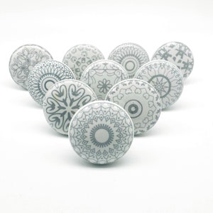 May include: A collection of eleven white ceramic cabinet knobs with various gray floral and geometric patterns. The knobs are arranged in a triangular shape against a white background, showcasing their decorative designs.