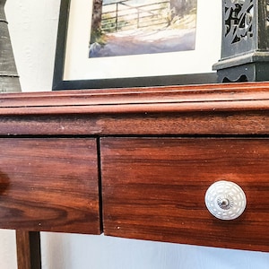 Op de afbeelding: Een houten tafel met twee laden en witte keramische knoppen. De tafel heeft een donkerbruine afwerking en is gemaakt van massief hout.