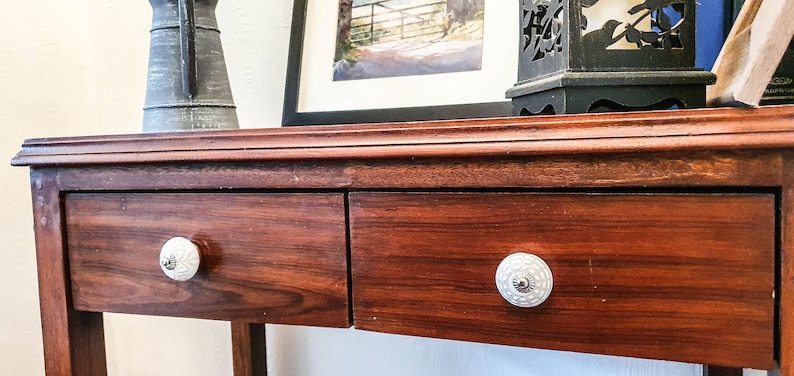 Op de afbeelding: Een houten tafel met twee laden en witte keramische knoppen. De tafel heeft een donkerbruine afwerking en is gemaakt van massief hout.
