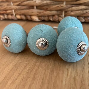 Puede incluir: Un conjunto de cinco pomos de armario azul claro con superficie texturizada y herrajes plateados. Los pomos est&aacute;n dispuestos sobre una superficie de madera, con una cesta tejida en el fondo.
