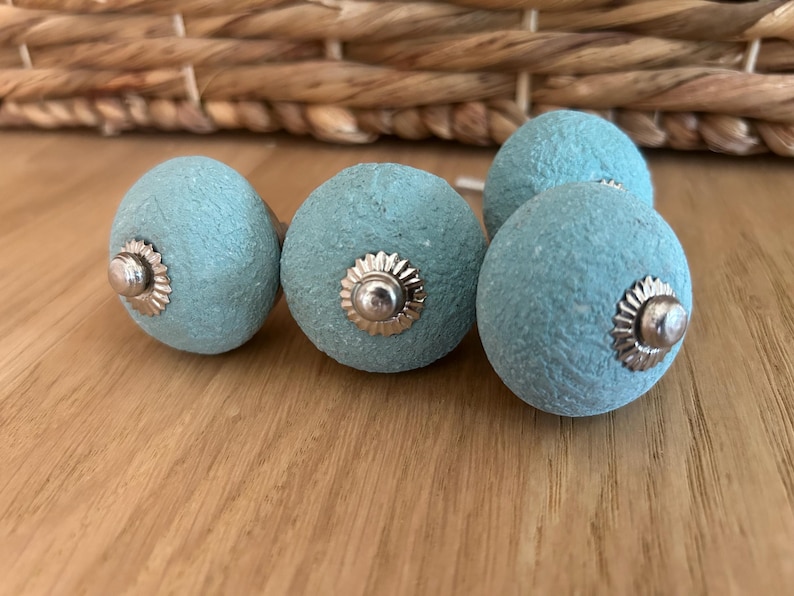 Puede incluir: Un conjunto de cinco pomos de armario azul claro con superficie texturizada y herrajes plateados. Los pomos est&aacute;n dispuestos sobre una superficie de madera, con una cesta tejida en el fondo.