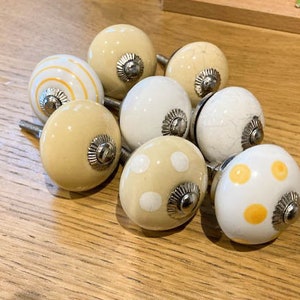 May include: A selection of eight ceramic cabinet knobs in cream and white. Some knobs feature stripes, polka dots, and a crackle effect. Each knob has a silver-coloured metal base and is designed for furniture.