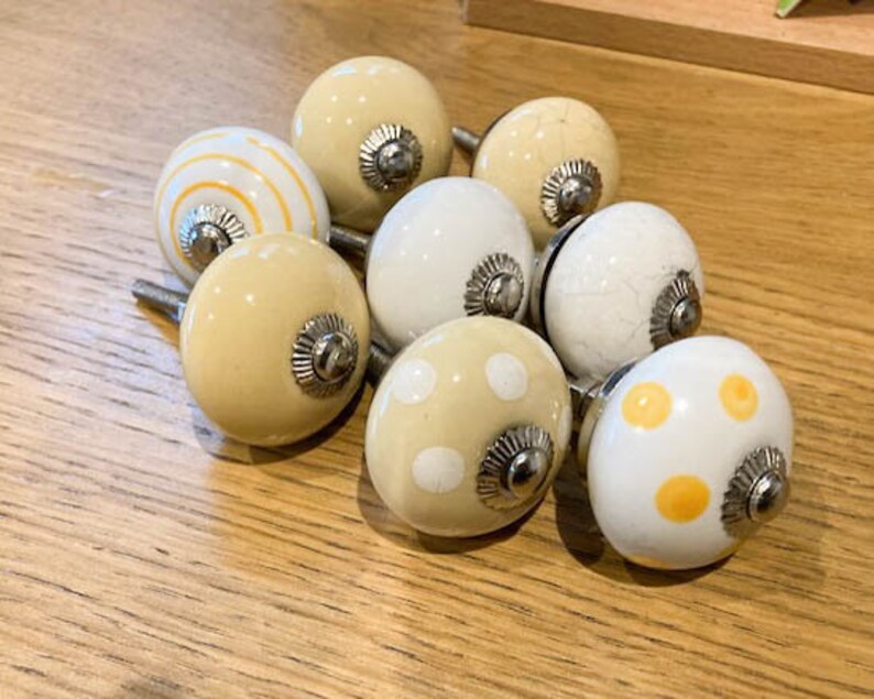May include: A selection of eight ceramic cabinet knobs in cream and white. Some knobs feature stripes, polka dots, and a crackle effect. Each knob has a silver-coloured metal base and is designed for furniture.