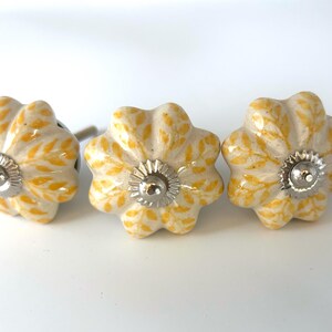 May include: Three flower-shaped cabinet knobs. The knobs are white with yellow leaf patterns and silver metal hardware. The knobs are ceramic and have a glossy finish. These decorative knobs are ideal for furniture.
