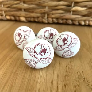 Op de afbeelding: Vier witte keramische kastknoppen met een bordeauxrood bloemmotief. De knoppen zijn rond en hebben een gedetailleerd bloemen- en bladerpatroon. Ze zijn gerangschikt op een houten oppervlak, met een gevlochten mand op de achtergrond.