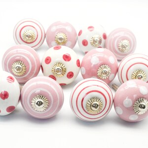 May include: A set of 12 pink and white ceramic knobs with red polka dots, stripes, and solid colors. The knobs have a gold accent in the center.