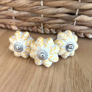 May include: Three flower-shaped cabinet knobs. The knobs are white with yellow leaf patterns and silver metal centres. They are arranged on a wooden surface, with a woven basket in the background.