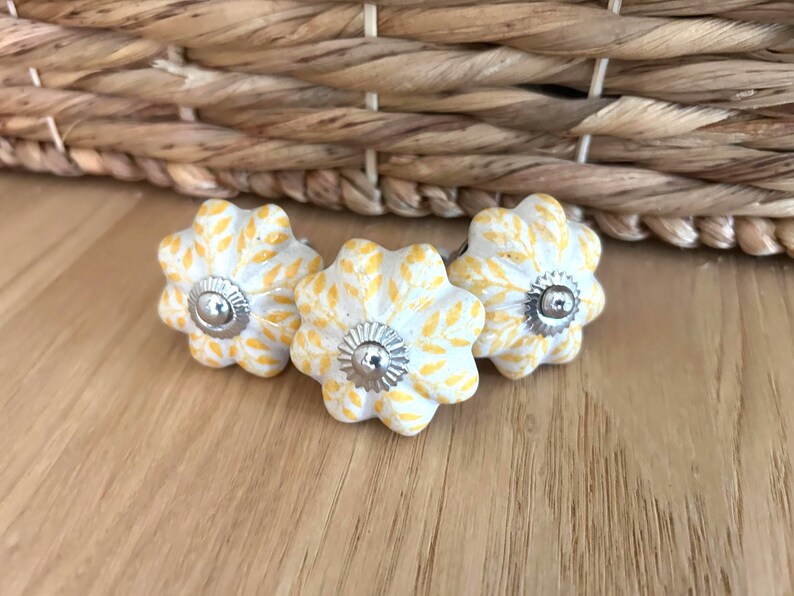 May include: Three flower-shaped cabinet knobs. The knobs are white with yellow leaf patterns and silver metal centres. They are arranged on a wooden surface, with a woven basket in the background.
