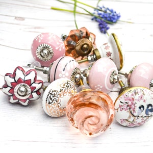 May include: Assortment of pink and white ceramic cabinet knobs with floral and geometric designs. The knobs have a vintage style and are perfect for adding a touch of charm to any home.