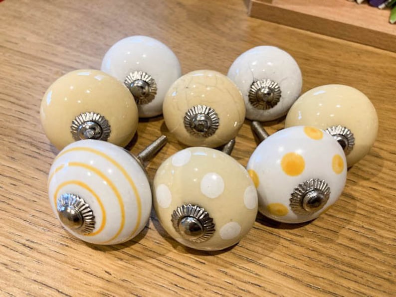 May include: Eight decorative cabinet knobs in cream and white. Some knobs have yellow stripes or polka dots. Each knob has a silver-coloured base and a metal screw for attachment. The knobs are arranged on a wooden surface, suitable for furniture or decor.