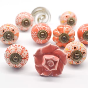 May include: Assortment of ten ceramic drawer knobs in various floral and geometric designs. The knobs are in shades of peach, coral, and white with gold accents.