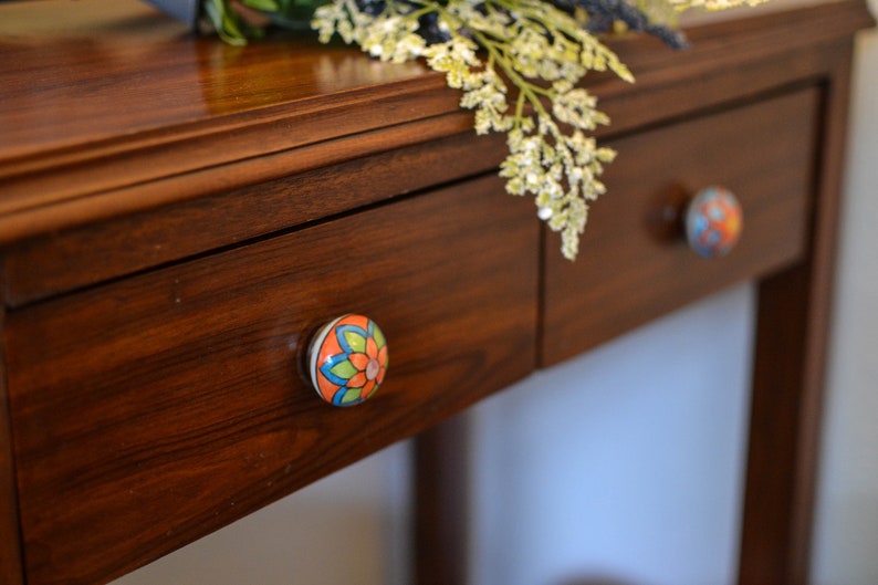 以下が含まれることがあります： 花柄のセラミックノブが付いた木製の引き出しが2つ。引き出しは濃い木製のテーブルの上にあります。