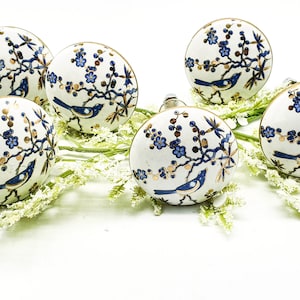 Puede incluir: Seis pomos de armario de cerámica blanca con borde dorado. Cada pomo presenta un pájaro azul posado en una rama dorada con flores azules.