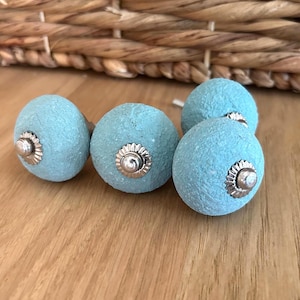 Könnte beinhalten: Fünf hellblaue Schrankknöpfe mit silberfarbenen Metallakzenten. Die Knöpfe haben ein strukturiertes, kugelförmiges Design. Die Knöpfe werden auf einer Holzoberfläche präsentiert, was auf ein Thema für Heimdekoration oder Hardware hindeutet.