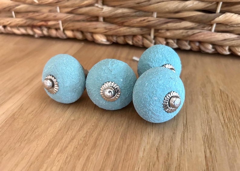 Puede incluir: Cinco pomos de armario azul claro con detalles met&aacute;licos plateados. Los pomos tienen un dise&ntilde;o esf&eacute;rico texturizado. Los pomos se muestran sobre una superficie de madera, lo que sugiere un tema de decoraci&oacute;n del hogar o ferreter&iacute;a.