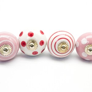 May include: Set of four ceramic drawer knobs with pink and red polka dot and stripe patterns. The knobs have a gold finish.
