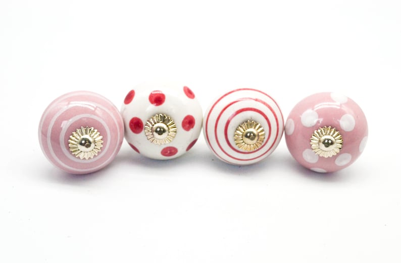 May include: Set of four ceramic drawer knobs with pink and red polka dot and stripe patterns. The knobs have a gold finish.