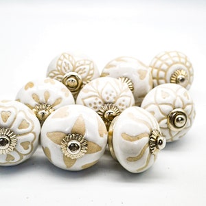 Op de afbeelding: Een verzameling van acht keramische knoppen met een witte basis en beige bloemmotieven. De knoppen hebben een gouden metalen accent in het midden.
