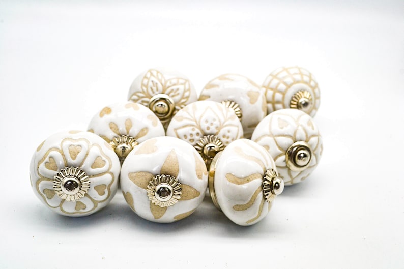 Op de afbeelding: Een verzameling van acht keramische knoppen met een witte basis en beige bloemmotieven. De knoppen hebben een gouden metalen accent in het midden.