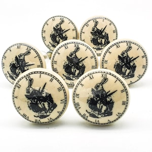 Puede incluir: Conjunto de siete pomos de armario de cerámica blanca con diseño de conejo negro y esfera de reloj. Cada pomo presenta un conejo negro sosteniendo un reloj de bolsillo con números romanos.