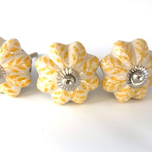 May include: Three flower-shaped cabinet knobs. The knobs are off-white with a yellow floral pattern and silver-coloured metal hardware. The knobs are arranged in a row against a white background.