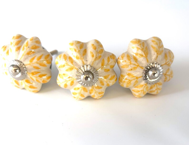 May include: Three flower-shaped cabinet knobs. The knobs are off-white with a yellow floral pattern and silver-coloured metal hardware. The knobs are arranged in a row against a white background.