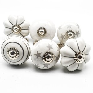 以下が含まれることがあります： 銀色のアクセントが付いた白いセラミック製のキャビネットノブ6個セット。ノブは、ストライプ、星、波状の縁など、さまざまなデザインが施されています。