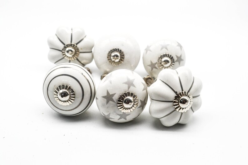 以下が含まれることがあります： 銀色のアクセントが付いた白いセラミック製のキャビネットノブ6個セット。ノブは、ストライプ、星、波状の縁など、さまざまなデザインが施されています。