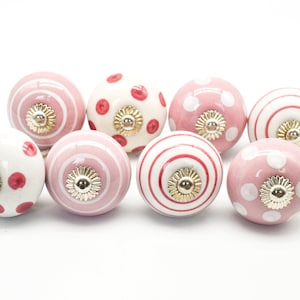 May include: Set of eight pink and white ceramic cabinet knobs with different patterns, including polka dots, stripes and swirls. Each knob has a gold-coloured accent.