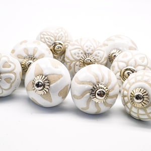 Op de afbeelding: Tien witte keramische kastknoppen met een beige bloemenpatroon. Elke knop heeft een gouden metalen accent in het midden.