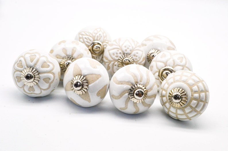 Op de afbeelding: Tien witte keramische kastknoppen met een beige bloemenpatroon. Elke knop heeft een gouden metalen accent in het midden.