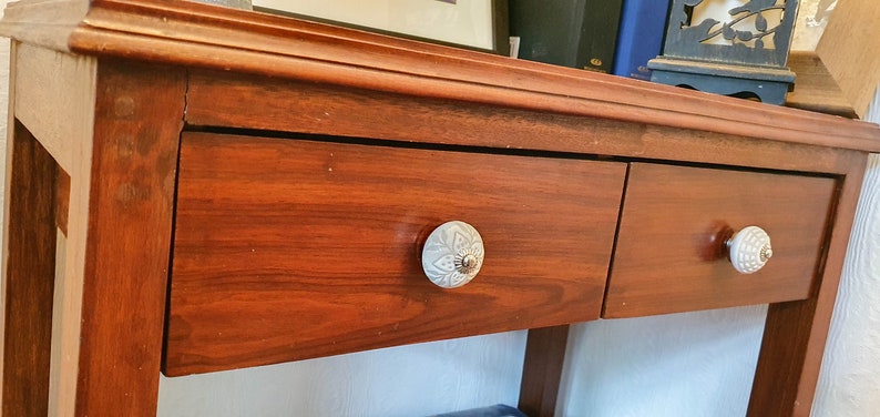 Op de afbeelding: Een houten consoletafel met twee laden. De laden hebben decoratieve witte keramische knoppen. De tafel is donker roodbruin.