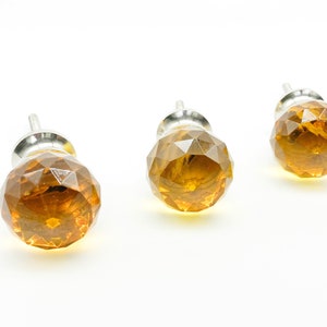 May include: Three amber-colored faceted glass cabinet knobs with silver-toned metal bases. The knobs are arranged on a white background, showcasing their geometric design and warm color. These decorative knobs are suitable for furniture upgrades.