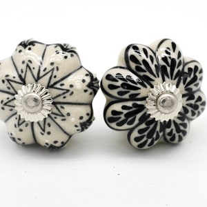 May include: Two ceramic flower-shaped cabinet knobs. The knobs are off-white with black floral designs and silver-coloured metal hardware. The knobs are decorative and suitable for furniture.