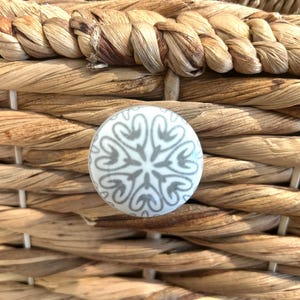 Puede incluir: Primer plano de una cesta tejida con un pomo decorativo redondo. El pomo es blanco con un diseño floral gris. La cesta está hecha de material marrón claro, tejido apretado.