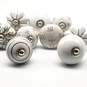 以下が含まれることがあります： 銀色のアクセントが付いた白いセラミック製のキャビネットノブ8個セット。各ノブは、星、雪片、時計の文字盤など、異なるデザインが施されています。