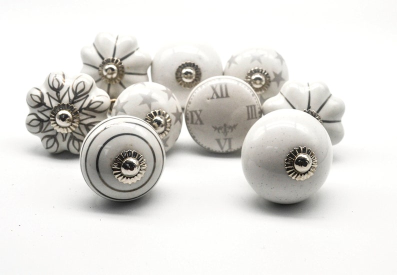 以下が含まれることがあります： 銀色のアクセントが付いた白いセラミック製のキャビネットノブ8個セット。各ノブは、星、雪片、時計の文字盤など、異なるデザインが施されています。