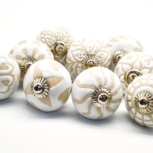 Op de afbeelding: Een verzameling van tien witte keramische kastknoppen met gouden accenten. Elke knop heeft een ander ingewikkeld ontwerp in beige.