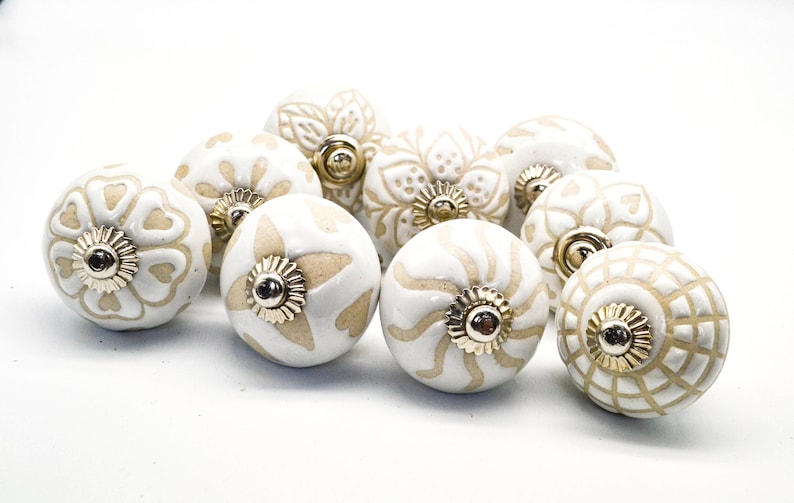 Op de afbeelding: Een verzameling van tien witte keramische kastknoppen met gouden accenten. Elke knop heeft een ander ingewikkeld ontwerp in beige.